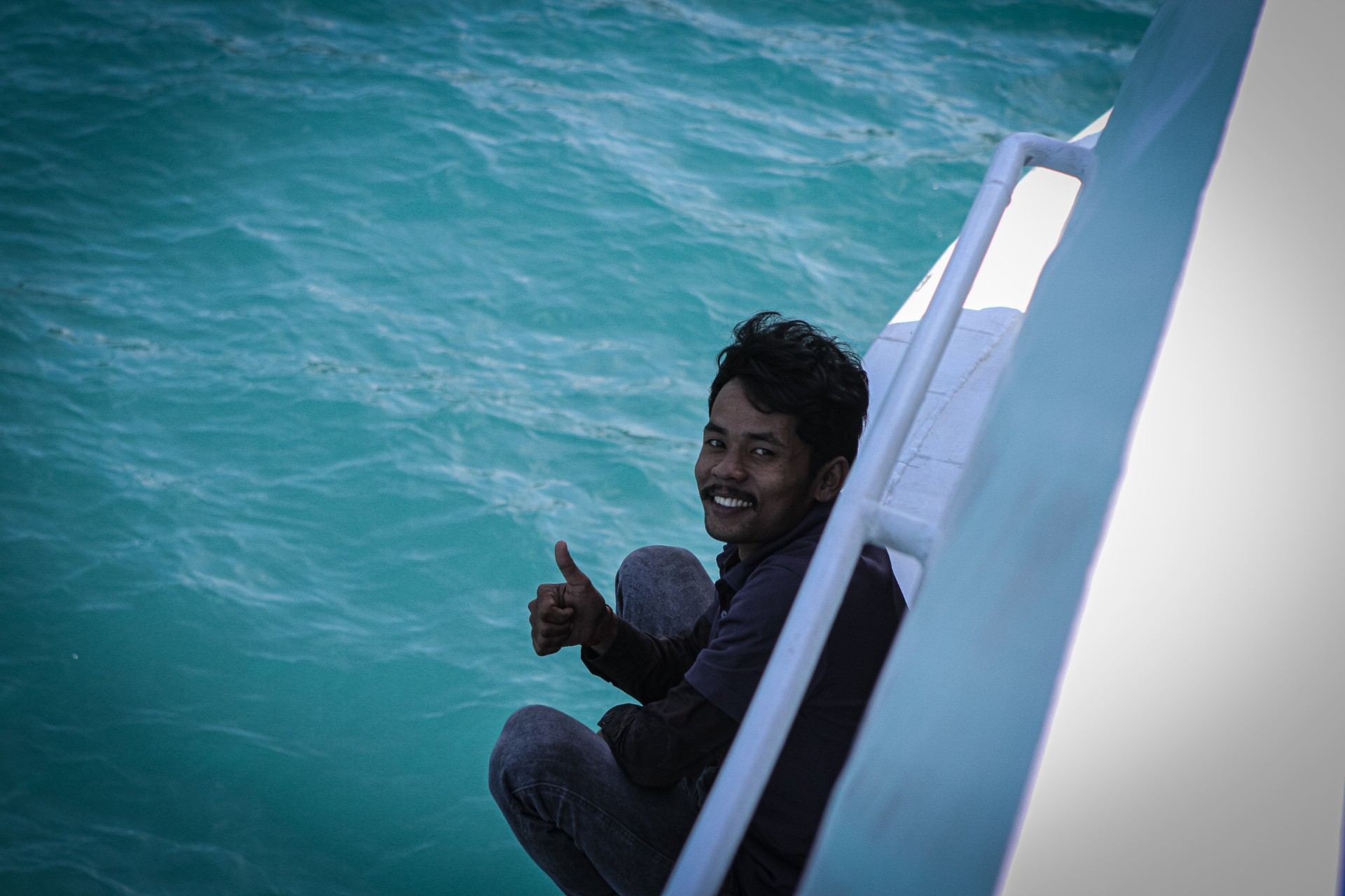 A man leans out of a boat over water, grinning at the camera and giving a thumbs up