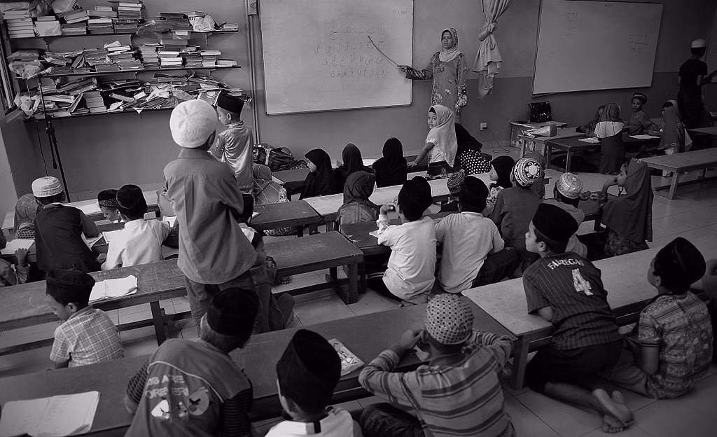 A woman in a hijab points to a whiteboard in front of a class on young children. One boy stands up, facing the board.