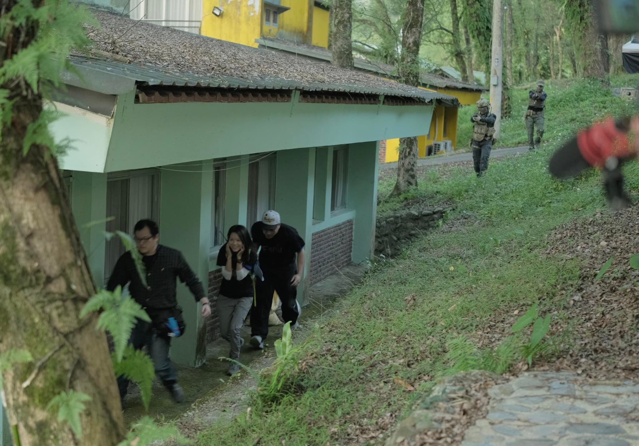Three people run along the side of a building, followed at a distance by two people wearing army gear.