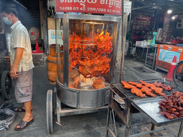 A vertical rack of cooked chickens are on the street next to a barbeque.