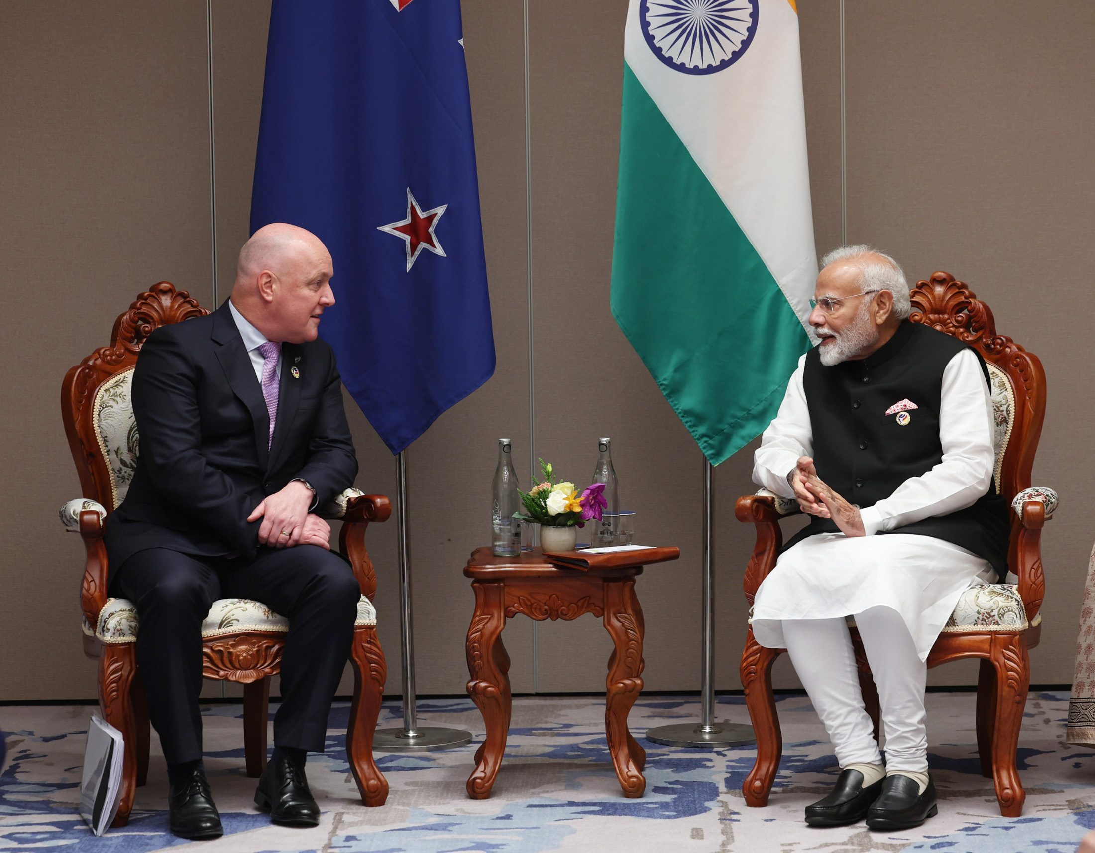 New Zealand Prime Minister Christopher Luxon sits in a chair and leans over towards Indian Prime Minister Narendra Modi. A flag from each respective country hangs in the background.
