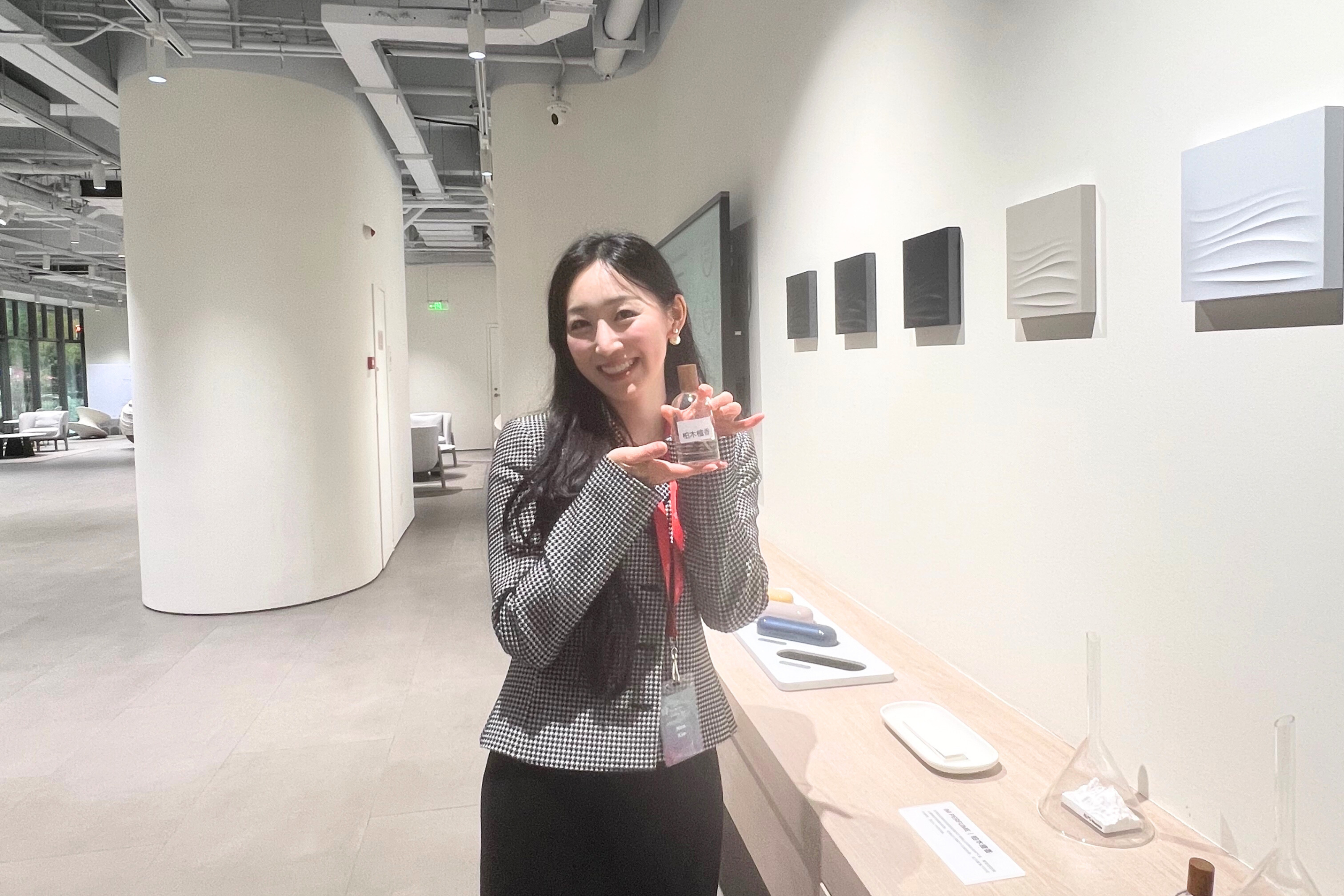 A woman stands in a white open space, smiling and holding a small bottle of perfume up to her face