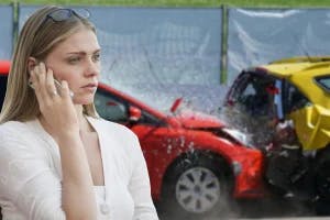 A woman calling the police with a car accident in the background.