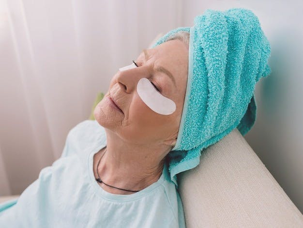 Senior woman reclining with eye pads post-eyelid surgery for swelling reduction.