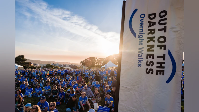 A crowd of people gathered in the sun next to a banner about the Overnight Walks