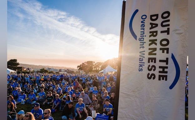 A crowd of people gathered in the sun next to a banner about the Overnight Walks