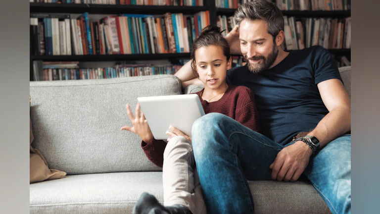 Father and daughter sitting on sofa looking at electronic tablet