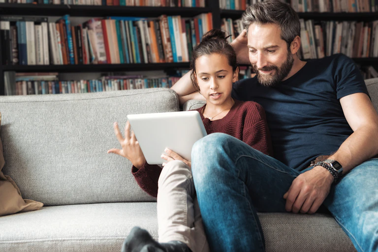 Father and daughter sitting on sofa looking at electronic tablet