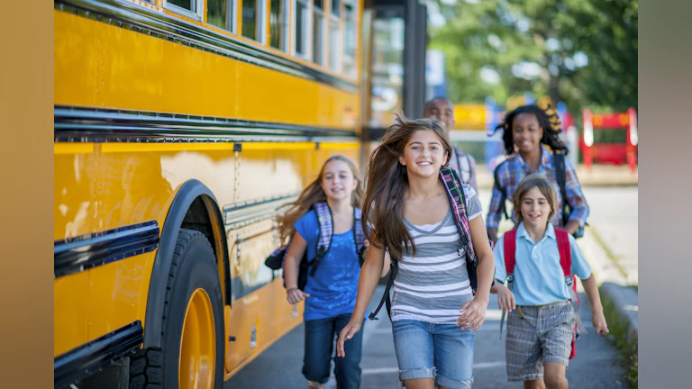 Children stepping out of a school bus, smiling and laughing as they run into school.