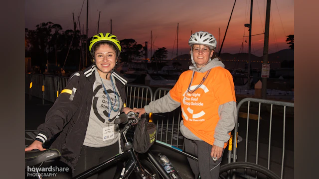 Two people in bike helmets and AFSP shirts standing beside bikes pose in front of a sunset.