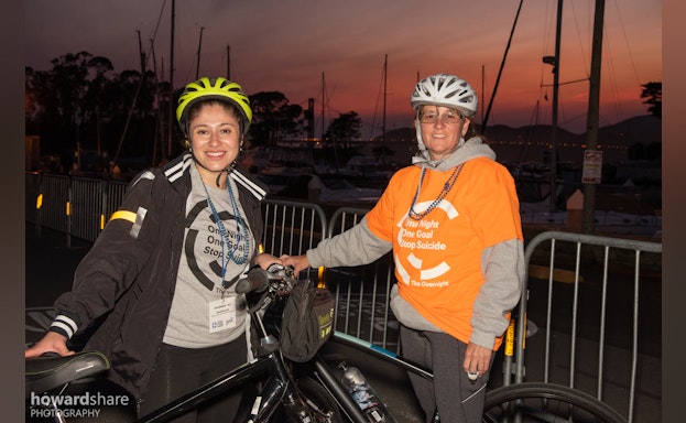Two people in bike helmets and AFSP shirts standing beside bikes pose in front of a sunset.