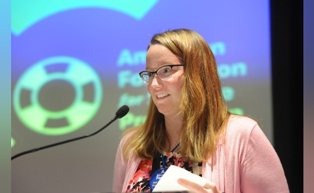 Jen giving a speech at a microphone in front of a screen that reads: "American Foundation for Suicide Prevention"