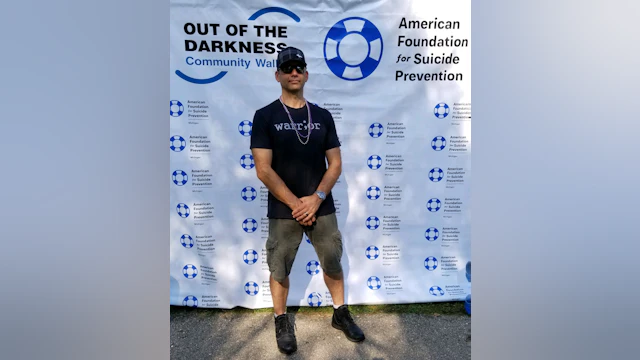 Ian Perry standing with his arms crossed in front of an Out of the Darkness community walk poster