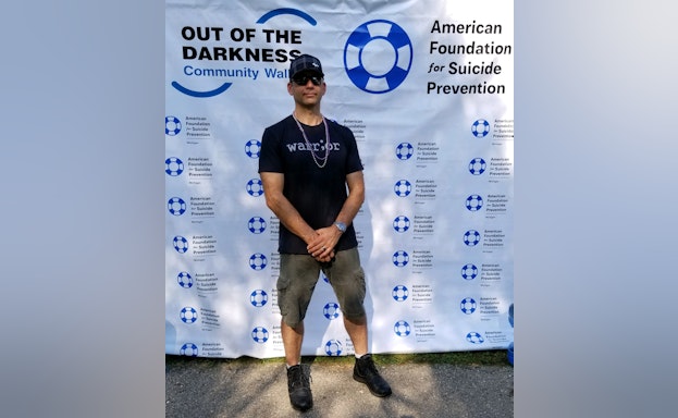 Ian Perry standing with his arms crossed in front of an Out of the Darkness community walk poster