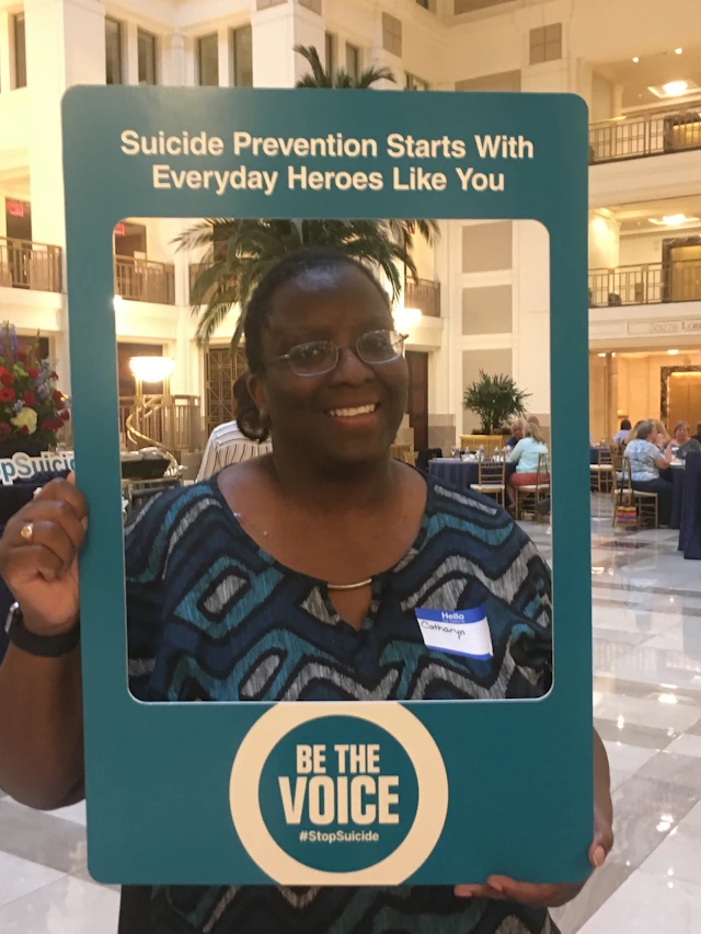 Catharyn Turner posing with a "Be The Voice" photo frame that says, "Suicide Prevention Starts With Everyday Heroes Like You"
