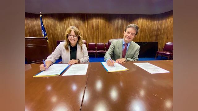 Maggie and Douglas smile at the camera while holding pens and signing the agreement