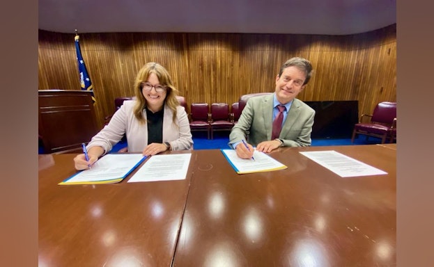 Maggie and Douglas smile at the camera while holding pens and signing the agreement