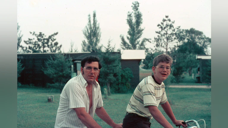 Father and son riding bikes together with a lawn and trees in the background.