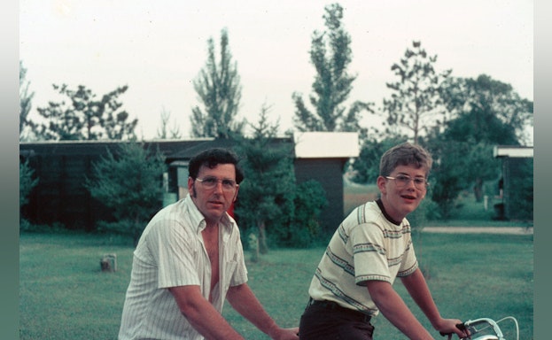 Father and son riding bikes together with a lawn and trees in the background.