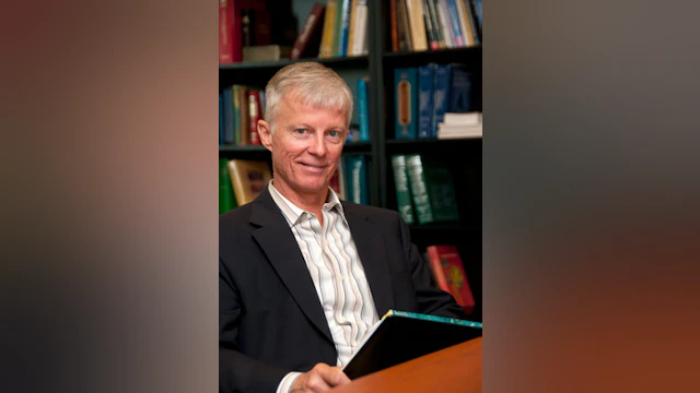 Gregory Ordway smiling while sitting at a wooden table with bookshelves in the background.