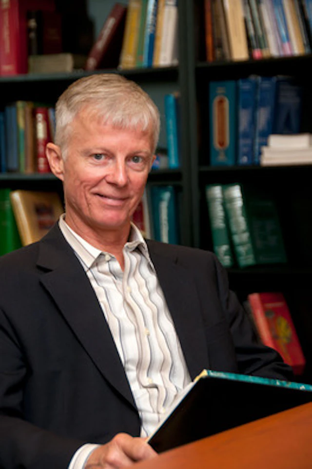 Gregory Ordway smiling while sitting at a wooden table with bookshelves in the background.