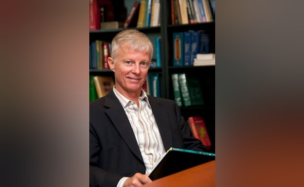 Gregory Ordway smiling while sitting at a wooden table with bookshelves in the background.