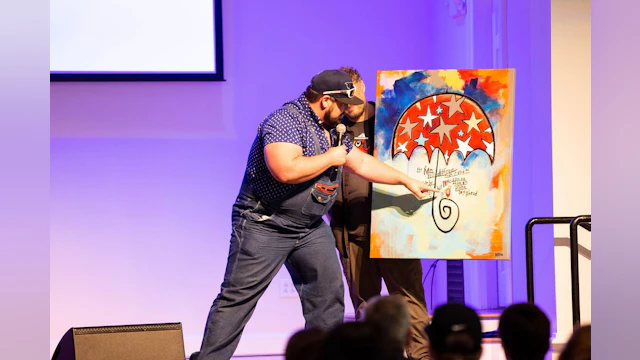 A man holds a microphone and points to a painting in a silent auction