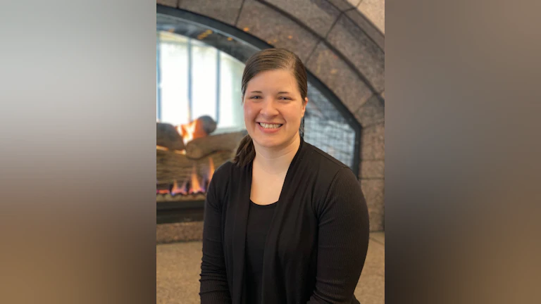 The author, Tarra Grammenos, wearing a black top and cardigan, smiling and sitting in front of a fireplace.