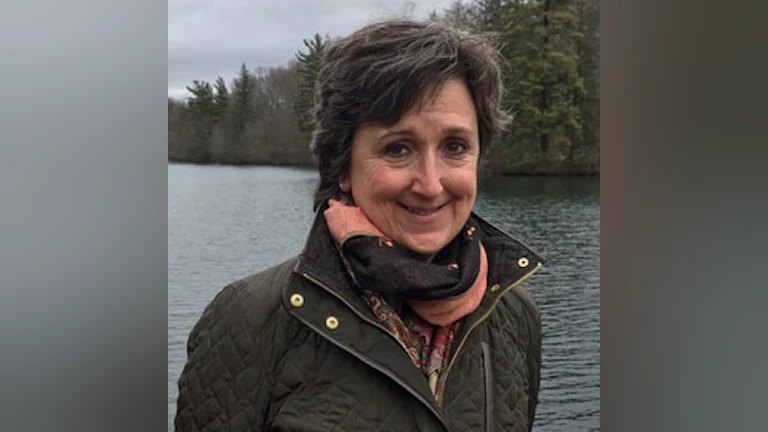 The author, Meg Kissinger, wearing a scarf and dark green jacket. She is smiling and standing in front of a lake with coniferous trees along the shoreline.