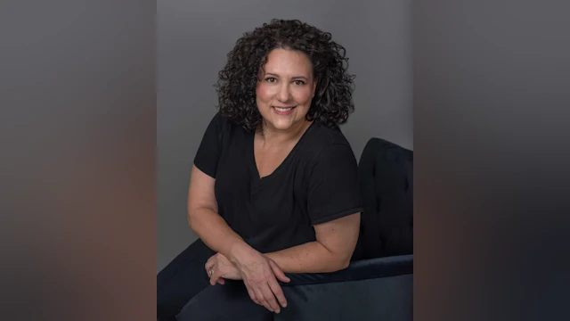 Karen Carreira smiling while sitting in a chair against a dark grey backdrop.
