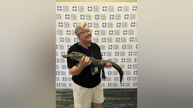 Kris Williams smiling and holding a reptile, with AFSP lifesaver logos on a backdrop behind her.