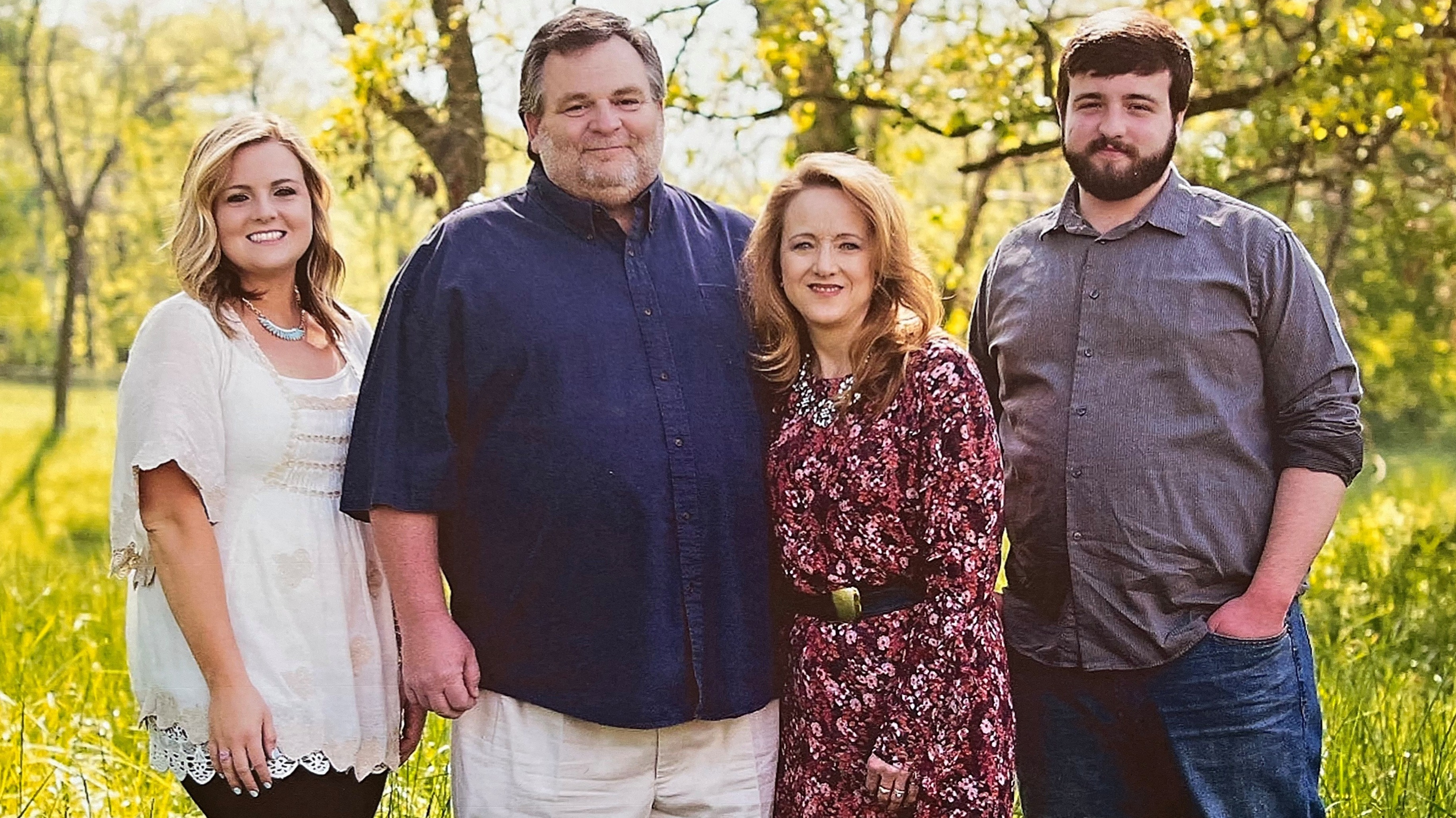 The Kerby family smiling together for a photo. They're standing outside on a beautiful, green, and sunny day.