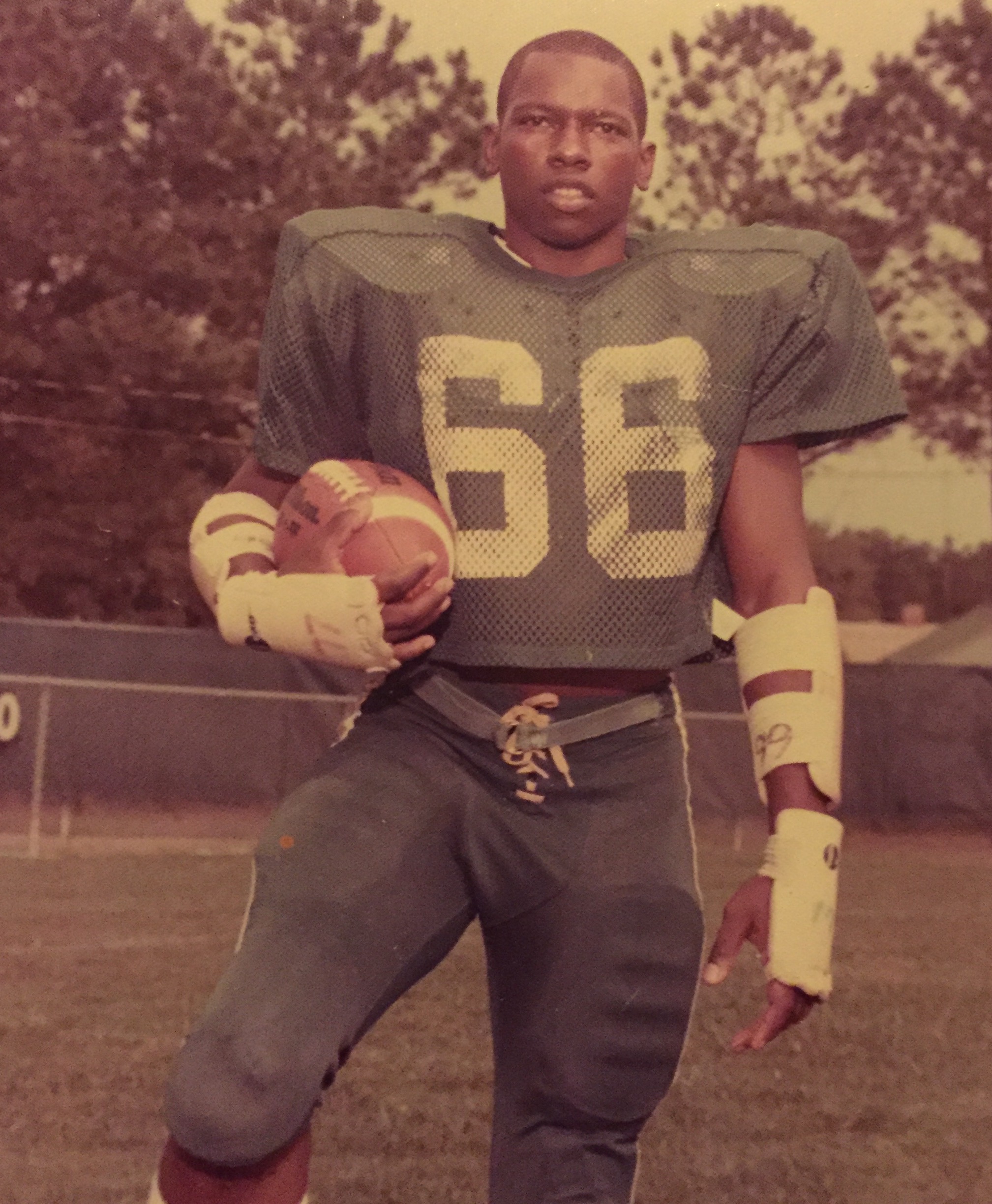 Vic pictured as a teenager, posing for a portrait on a sports field. He's wearing a uniform and holding a football.
