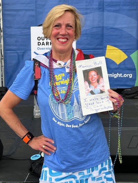 The author, Ellen Marks, walking in honor of her late wife Monica Boll at the Overnight. She's smiling, wearing a blue AFSP t-shirt, and holding a picture of Monica.
