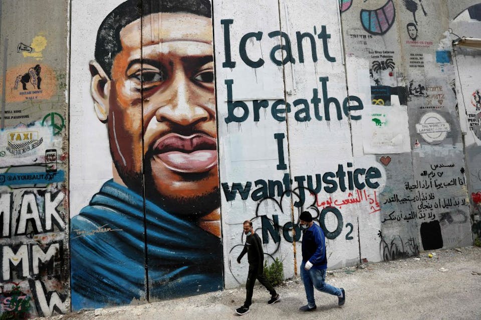 People walk past a mural of George Floyd on March 31, 2021, painted on a section of Israel's separation barrier in the city of Bethlehem. *Photo by Emmanuel Dunand/AFP via Getty Images.* People walk past a mural showing the face of George Floyd, an unarmed handcuffed black man who died after a white policeman knelt on his neck during an arrest in the US, painted on a section of Israel’s controversial separation barrier in the city of Bethlehem in the occupied West Bank on March 31, 2021. – The teenager who took the viral video of George Floyd’s death said on March 30, at the trial of the white police officer charged with killing the 46-year-old Black man that she knew at the time “it wasn’t right.” Darnella Frazier, 18, was among the witnesses who gave emotional testimony on Tuesday at the high-profile trial of former Minneapolis police officer Derek Chauvin. Chauvin, 45, is charged with murder and manslaughter for his role in Floyd’s May 25, 2020 death, which was captured on video by Frazier and seen by millions, sparking anti-racism protests around the globe. (Photo by Emmanuel DUNAND / AFP) (Photo by EMMANUEL DUNAND/AFP via Getty Images)