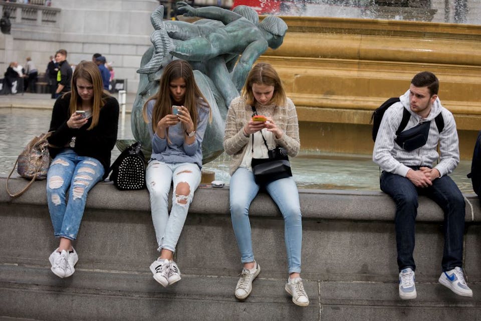 In London, three teenage girls are intently focused on their smartphones while a young man looks on. *Photo by In Pictures Ltd./Corbis via Getty Images. * Clifford-Main