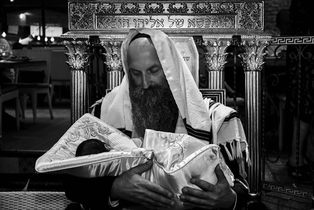 Orthodox Jewish man holding a newborn baby wrapped in a ceremonial blanket during the brit milah circumcision ceremony (Shutterstock)