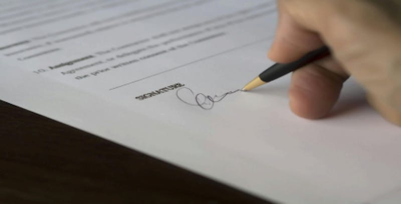 The image shows a close-up of a person signing a document. The person's hand is holding a black and gold pen, and they are writing their signature on the designated signature line at the bottom of the paper. The document includes some text, but the details are blurred and illegible.