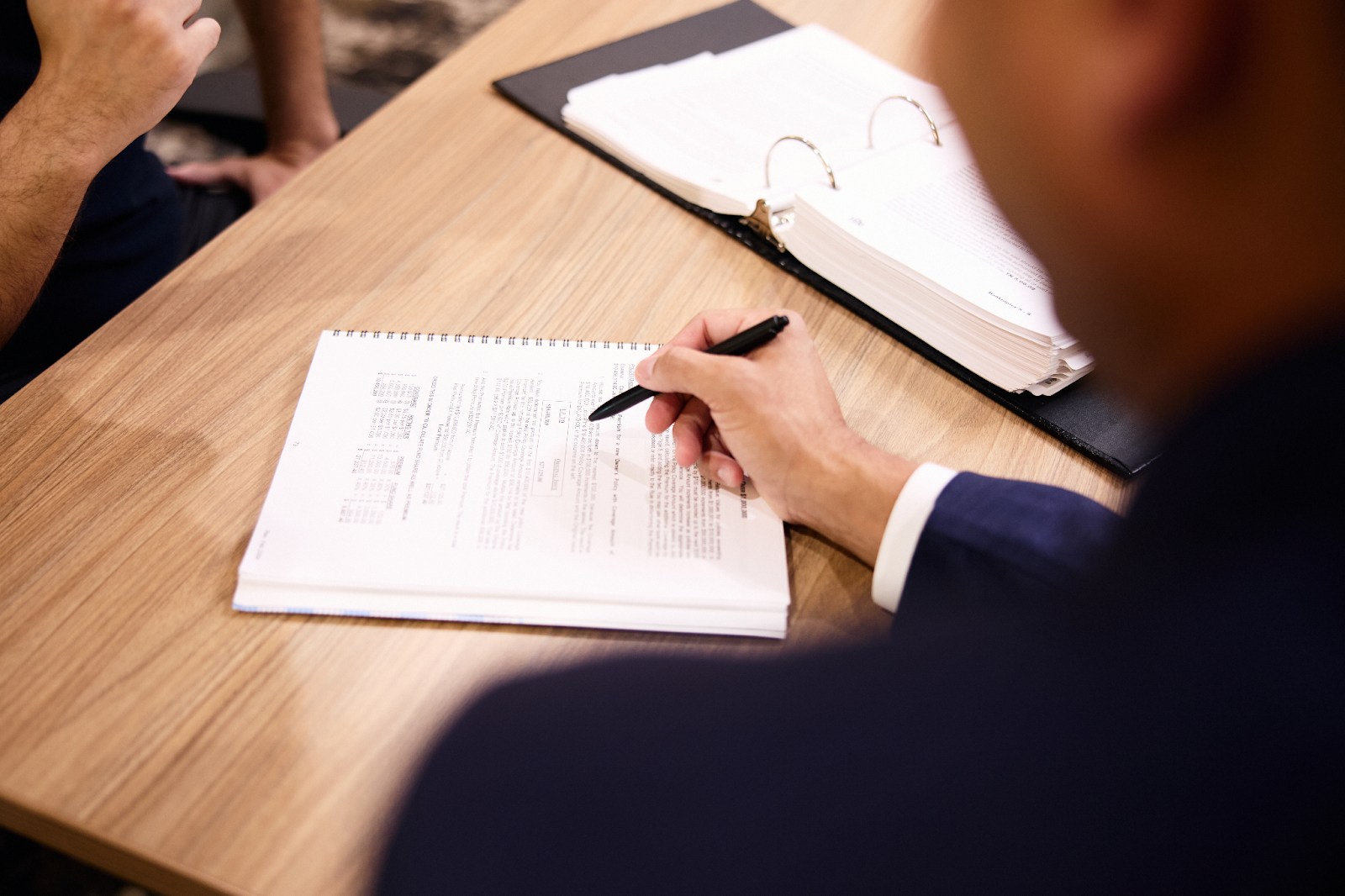 A person in a suit is writing on a document at a wooden desk, with another individual across from them. An open binder with papers suggests a business or legal meeting. The focus is on the hand holding the pen. 