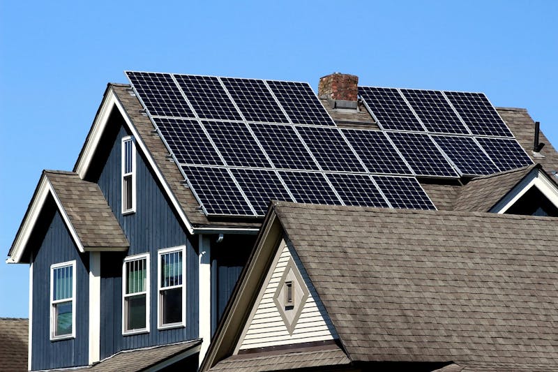 This image shows a house with solar panels on the roof. The house is blue with white trim, and the solar panels are lined up neatly to capture sunlight for energy. The sky is clear and blue.