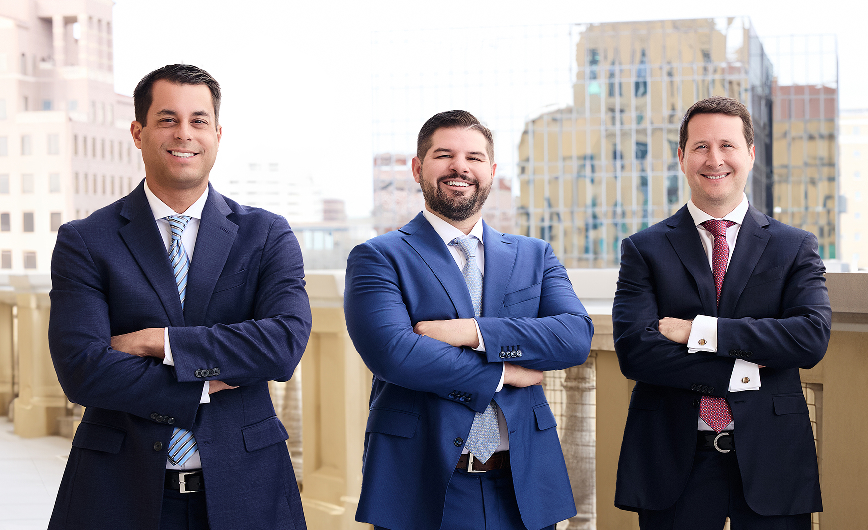Partners of Vargas Gonzalez Delombard LLP—Rene Delombard (left), Louis Gonzalez (middle), and Andrew Vargas (right)—standing outdoors with a cityscape in the background, dressed in business suits and smiling.