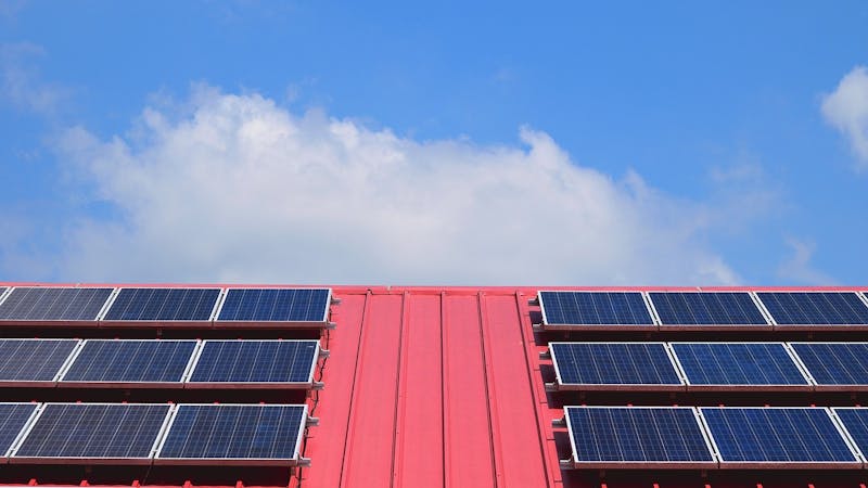 A close-up view of solar panels mounted on a bright red metal roof, under a blue sky with a few white clouds.