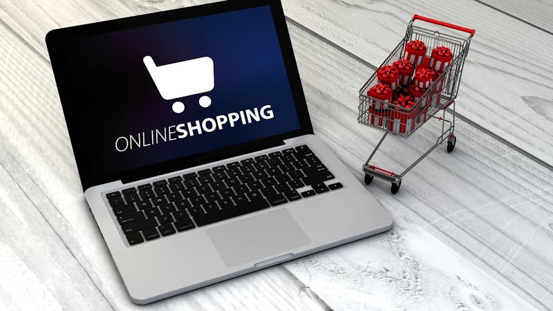 Laptop displaying 'Online Shopping' next to a miniature shopping cart filled with wrapped gift boxes on a wooden surface