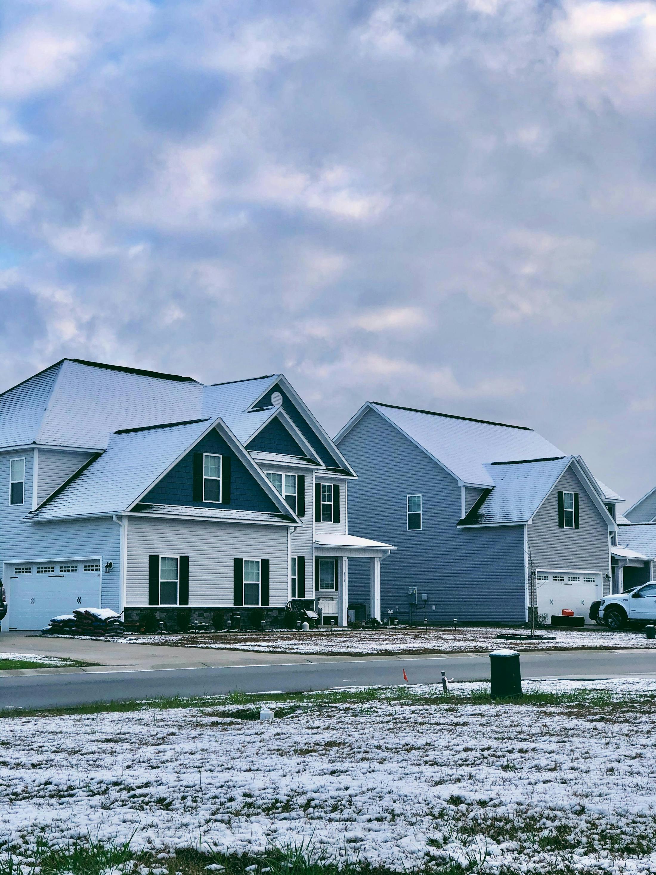 A row of houses with snow covered roofs and lawns. The houses are two-story with attached garages and front porches. The sky is cloudy.