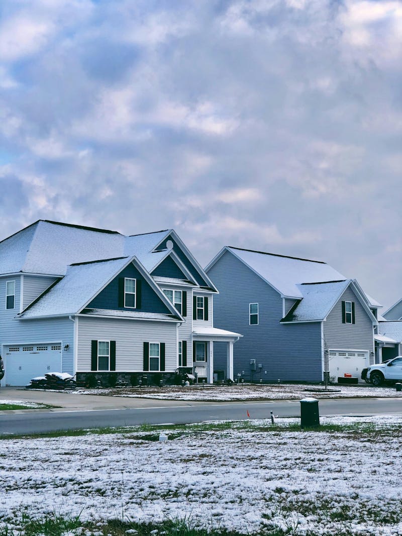 A row of houses with snow covered roofs and lawns. The houses are two-story with attached garages and front porches. The sky is cloudy.