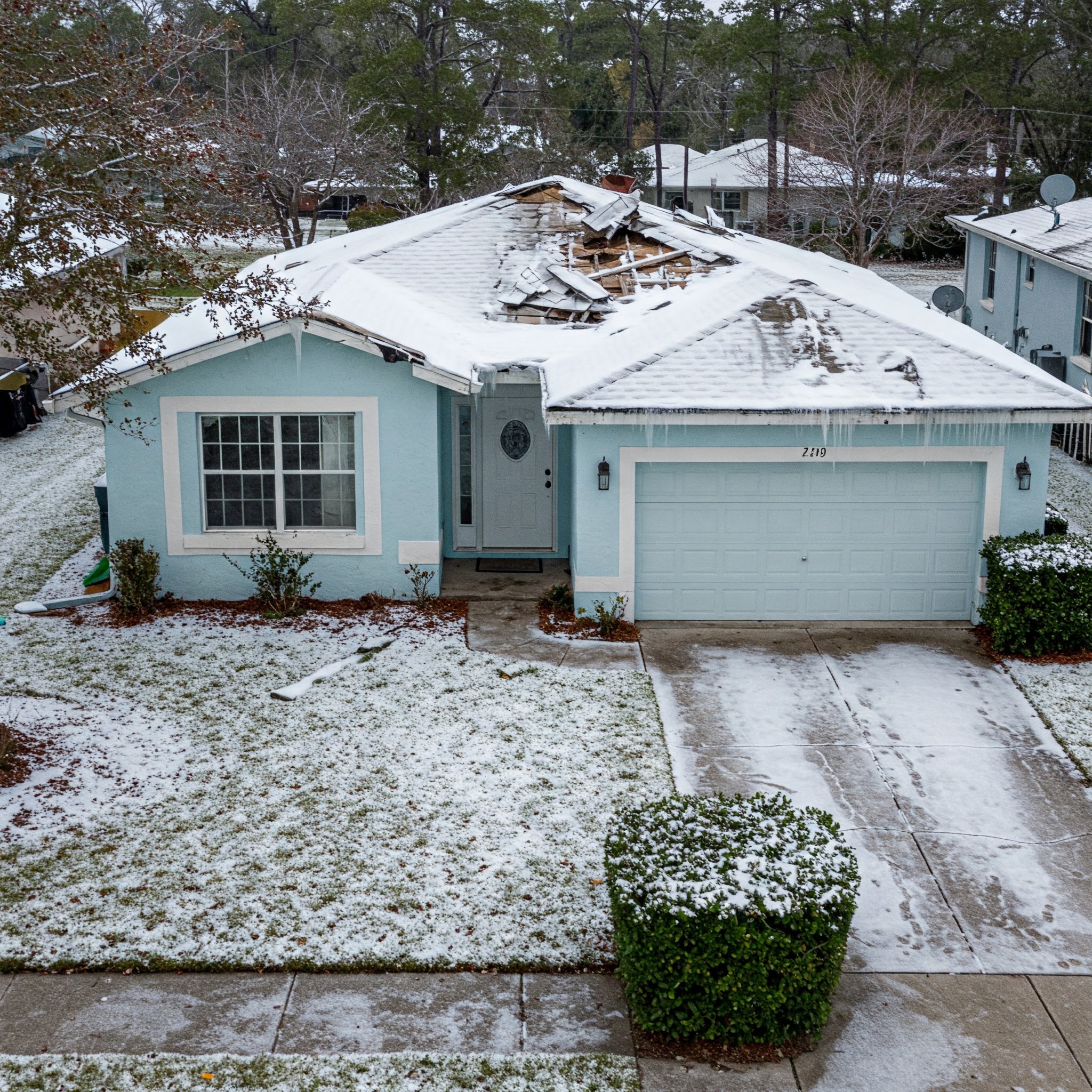 A blue, single-story house with roof damage. A large portion of the roof is missing, with shingles scattered across the lawn. The house is covered in snow, and there are icicles hanging from the gutters.