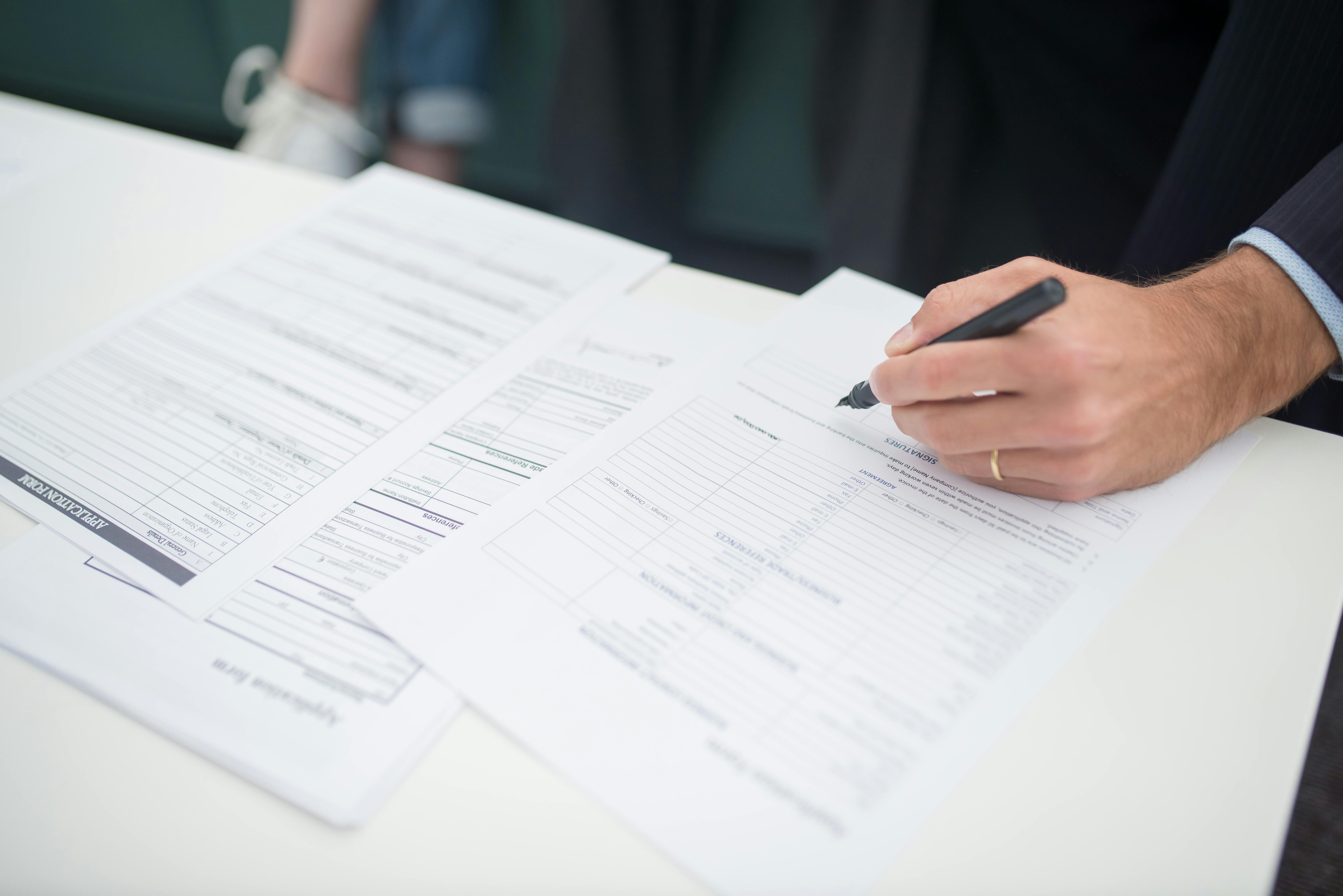 A person in a suit holding a pen and filling out paperwork on a white desk, with multiple documents spread out.