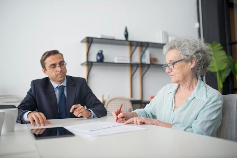 An older woman with gray hair and glasses is signing a document with a red pen, while a man in a suit, sits across from her observing. They are in a modern office setting with shelves in the background.