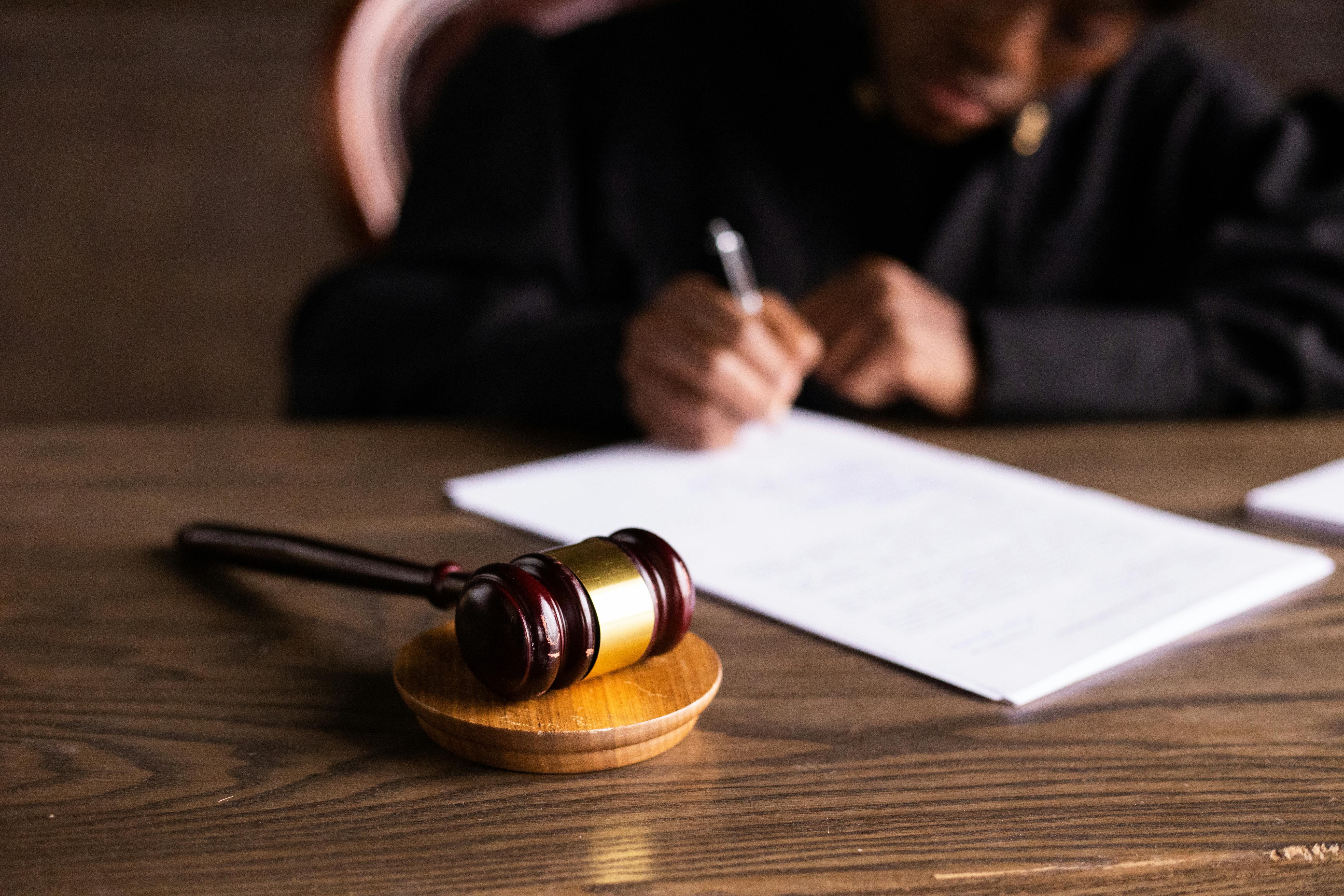 A wooden gavel rests on a table, while a judge in a black robe is seen in the background signing a document.