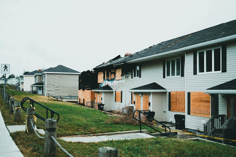 A row of houses with visible fire and structural damage, some windows boarded up, and caution tape surrounding the area.